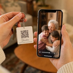 Op de afbeelding: Een witte vierkante sleutelhanger met een zwarte QR-code, het woord Grandma en de tekst SCAN TO WATCH OUR MEMORIES. Een smartphone toont een video van een oudere vrouw die twee kinderen knuffelt, passend bij de QR-code.