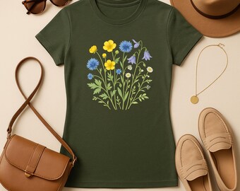 May include: Dark green t-shirt featuring a floral design with yellow, blue, and white flowers. The shirt is surrounded by accessories, including sunglasses, a purse, a hat, a necklace, and shoes. The t-shirt is a casual wear item.