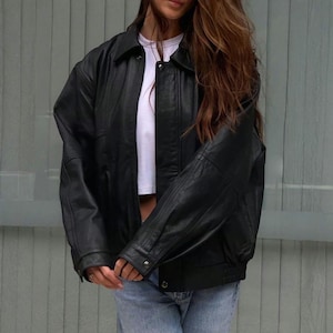 Peut inclure: Veste en cuir noire avec un col et des poignets boutonnés. La veste a une coupe décontractée et est portée sur un haut blanc et un jean bleu. La veste a une fermeture boutonnée sur le devant et un ourlet côtelé.