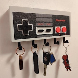 Könnte beinhalten: Ein weiß-grauer Schlüsselhalter im Design eines Nintendo-Controllers mit schwarzen Knöpfen und roten Akzenten. Der Schlüsselhalter hat fünf schwarze Haken und hält verschiedene Schlüssel und Schlüsselanhänger, darunter eine Deadpool-Figur.