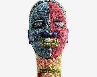 May include: A beaded mask sculpture with a split design. One half is blue, the other red. The mask has white, yellow, and blue accents. The neck is a cylinder of orange, yellow, and brown beads. The mask is a decorative art piece.