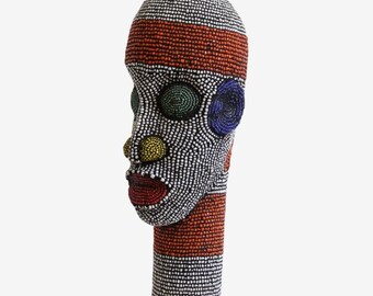 May include: A beaded sculpture of a human head. The head is covered in white beads with accents of red, black, yellow, green, and blue beads. The lips are red. The sculpture has a cylindrical neck.