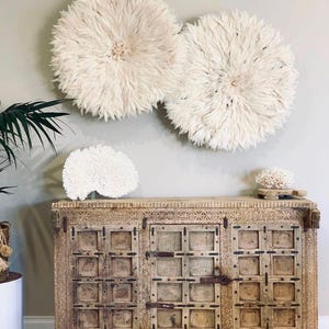 Peut inclure: Deux grandes décorations murales rondes de couleur crème avec une texture de plumes sont suspendues au-dessus d'un meuble en bois. Le meuble présente des détails sculptés et une quincaillerie en métal. Une sculpture blanche en forme de corail est posée sur le dessus.