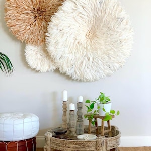 Peut inclure: Décor d'intérieur avec trois décorations murales rondes texturées, crème et beige. Une table en bois présente des bougies, une petite plante et des objets décoratifs. Un pouf blanc et marron est au premier plan.