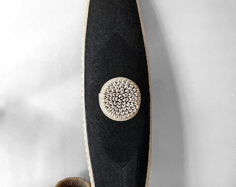 Puede incluir: Un gran escudo decorativo negro con una pieza central circular adornada con conchas. El escudo tiene un borde tejido y se exhibe junto a una pequeña cesta tejida. El escudo mide aproximadamente 152 cm de altura.