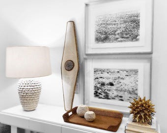 Puede incluir: Una consola blanca exhibe artículos de decoración del hogar. Una lámpara color crema con una base texturizada se encuentra junto a una pieza decorativa de madera alta. Dos impresiones de paisajes en blanco y negro enmarcadas cuelgan sobre la mesa, con una bandeja tejida y esferas decorativas.