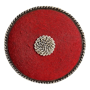 May include: A round, red beaded decorative item with a central cluster of white seashells. The edge is trimmed with small white seashells and a dark brown border. The item is circular and set against a white background.