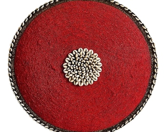 Puede incluir: Un objeto decorativo redondo de cuentas rojas con un grupo central de conchas blancas. El borde está adornado con pequeñas conchas blancas y un borde marrón oscuro. El artículo es circular y está sobre un fondo blanco.