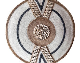 Puede incluir: Arte mural redondo con un diseño central de conchas marinas y cuerda. La obra de arte tiene forma circular con un fondo de cuentas blancas, acentuado por una franja azul oscuro y un borde marrón. Decorativo y texturizado.