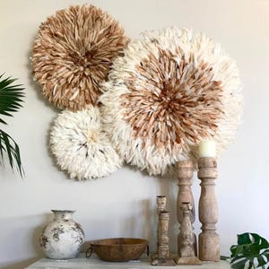 Peut inclure: Trois décorations murales rondes et texturées, dans des tons de blanc et de marron, sont accrochées à un mur clair. En dessous, une collection de bougeoirs en bois, un vase décoratif et un bol en bois sont disposés sur une surface blanche.