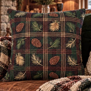 May include: A square throw pillow featuring a dark green, black, and red plaid pattern. The fabric is decorated with a repeating print of brown pinecones and green and gold pine needles scattered across the surface of the cushion.