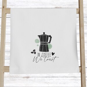 Może przedstawiać: Biały ręcznik kuchenny z czarną ilustracją kawiarki, ziaren kawy i napisem "In Coffee We Trust". Projekt zawiera zielone akcenty i symbol serca.