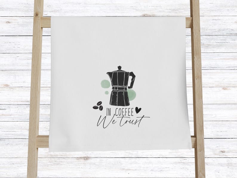 Może przedstawiać: Biały ręcznik kuchenny z czarną ilustracją kawiarki, ziaren kawy i napisem "In Coffee We Trust". Projekt zawiera zielone akcenty i symbol serca.