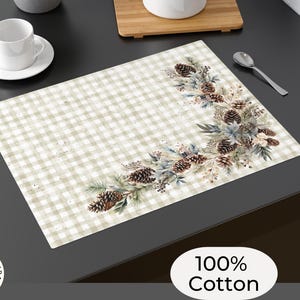 May include: A rectangular placemat with a beige and white checkered pattern. A decorative corner design features pine cones, blue-green leaves, and white flowers. The text "100% Cotton" is visible.