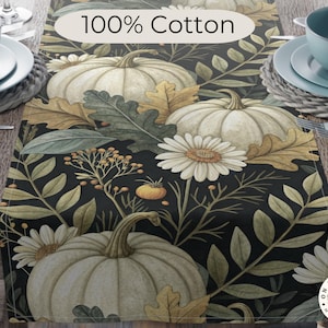 May include: A cotton table runner with a black background, featuring a pattern of white pumpkins, daisies, and autumn leaves. The text "100% Cotton" is displayed on the top. The runner is set on a wooden table, with tableware visible on either side.