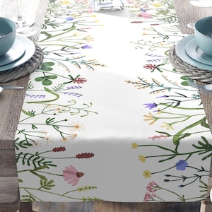 May include: White table runner with a floral design featuring wildflowers in shades of pink, yellow, purple, and green. The runner is set on a wooden table with blue bowls and plates.