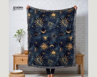 May include: A navy blue throw blanket featuring a celestial design with gold suns, moons, and stars. The blanket has a soft, fringed edge and is held up against a white brick wall. The brand name "gracious gifting co" is visible.