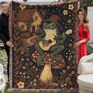 May include: Woven throw blanket featuring a whimsical illustration of a frog playing a guitar while seated on a mushroom. The design includes a house, flowers, and a path, with a fringed border. The color palette includes black, brown, green, and yellow.