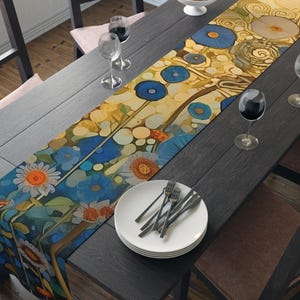 Puede incluir: Un camino de mesa floral con un diseño colorido con flores azules, amarillas y naranjas. El camino de mesa está colocado sobre una mesa de madera oscura, con platos, cubiertos y copas de vino. El tema general es la decoración del hogar.