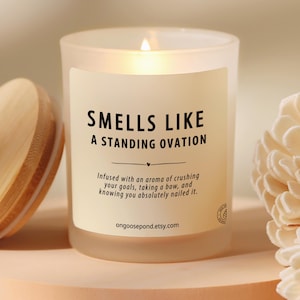 May include: A lit, frosted glass candle with the text "SMELLS LIKE A STANDING OVATION." The candle has a wooden lid and is next to a decorative flower. The candle's label includes the Etsy shop's URL.