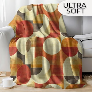 May include: Ultra soft blanket with a geometric pattern of large circles and shapes in shades of red, orange, beige, and brown. The blanket is draped over a white sofa, with the text "ULTRA SOFT" in white.