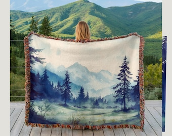 May include: A woven throw blanket featuring a watercolor landscape of a mountain forest in shades of blue and green. The blanket has a fringed edge and is held up against a backdrop of a mountain range.