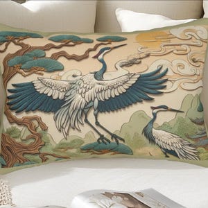May include: A rectangular throw pillow featuring a printed illustration of two cranes with white bodies and dark blue wings. The scene includes a stylised brown tree, green hills, and a yellow sun against a cream background with swirling clouds.