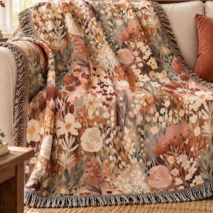 May include: A woven throw blanket featuring a dense botanical pattern of wildflowers and foliage in shades of rust, terracotta, cream, and muted green. The rectangular blanket has a decorative fringe along the edges and a textured finish.