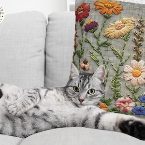 Pu&ograve; includere: Un gatto tabby argentato si rilassa su un divano grigio chiaro accanto a un cuscino decorativo. Il cuscino presenta un design floreale ricamato con fiori rossi, arancioni, gialli, viola, rosa e blu su sfondo grigio. Un articolo per la casa.