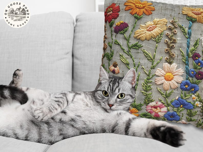 Pu&ograve; includere: Un gatto tabby argentato si rilassa su un divano grigio chiaro accanto a un cuscino decorativo. Il cuscino presenta un design floreale ricamato con fiori rossi, arancioni, gialli, viola, rosa e blu su sfondo grigio. Un articolo per la casa.