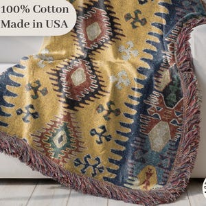 May include: A patterned throw blanket in yellow, navy, and burgundy, with a fringed edge, draped over a white sofa. The blanket features a geometric design. The text "100% Cotton Made in USA" is visible. A home decor item.
