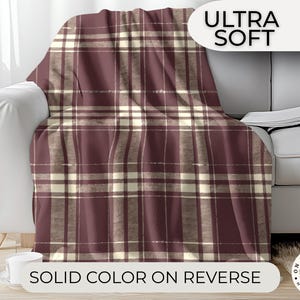 May include: A burgundy and cream plaid throw blanket draped over a white sofa. The blanket has a repeating pattern of intersecting lines. The words "ULTRA SOFT" and "SOLID COLOR ON REVERSE" are visible in the image.