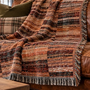 May include: A woven throw blanket featuring a multi-colored striped pattern in shades of rust, orange, brown, black, and cream. The rectangular textile has a textured, nubby surface and is finished with a fringed edge along the bottom border.