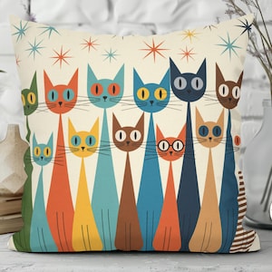 May include: Decorative throw pillow featuring a retro design of stylized cats in various colors, including orange, blue, and brown, against a cream background. The pillow has a mid-century modern aesthetic with starburst accents.