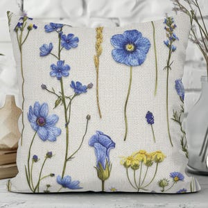 May include: Square throw pillow with a botanical design featuring blue and yellow wildflowers on a cream-colored background. The pillow has a textured appearance, with a variety of floral illustrations.
