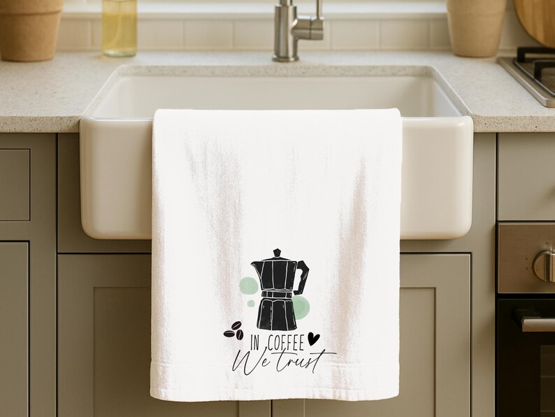 Może przedstawiać: Biały ręcznik kuchenny z czarną grafiką ekspresu do kawy, ziaren kawy i napisem "IN COFFEE WE TRUST". Ręcznik wisi nad białym zlewem.