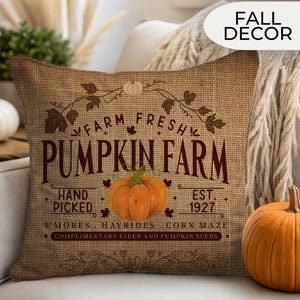 May include: A square burlap pillow with the text "FARM FRESH PUMPKIN FARM" and an orange pumpkin illustration. The pillow also has the text "HAND PICKED" and "EST. 1927". A decorative pumpkin sits next to the pillow.