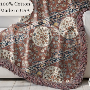 May include: A woven throw blanket with a vintage-inspired design, featuring a mix of rust, cream, and navy blue colors. The blanket has a fringed edge and is draped over a white sofa. The text "100% Cotton Made in USA" is visible.