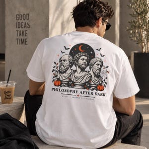 May include: A white t-shirt featuring a graphic on the back with three classical philosopher busts, pumpkins, bats, and a crescent moon. The text reads PHILOSOPHY AFTER DARK, STOIC, SPOOKY, TIMELESS. The design uses black, white, and orange colors.
