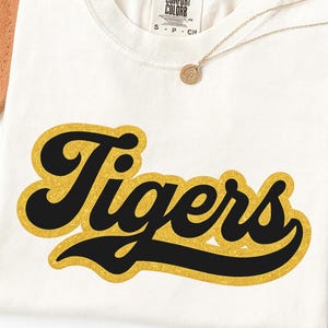 May include: An off-white short-sleeve t-shirt featuring the word Tigers in black script lettering with a gold glitter outline. The shirt has a crew neckline and a Comfort Colors brand label printed on the inside of the collar area.