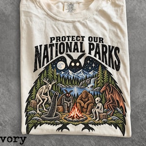 May include: An ivory t-shirt featuring a graphic print with the text "PROTECT OUR NATIONAL PARKS" above a scene of mythical creatures sitting around a campfire in a forest with mountains, a waterfall, and a large moth silhouette in the night sky.