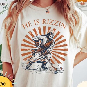 May include: Ivory-colored t-shirt with a graphic of Jesus playing hockey, halo above his head, and the text "HE IS RIZZIN". The design features brown and orange tones. The shirt has a round neck and short sleeves.