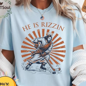 May include: Light blue t-shirt with the text "HE IS RIZZIN" above an illustration of Jesus playing hockey. The graphic features Jesus with a halo, hockey stick, and puck against a sunburst design. The shirt has a tag that says "%100 US COTTON".