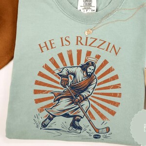 May include: A light green t-shirt with the text "HE IS RIZZIN" above a graphic of Jesus playing hockey. The graphic features Jesus with a halo, wearing a robe and hockey gear, set against a sunburst design. A gold necklace is draped on the shirt.