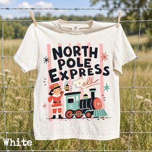 May include: A cream-colored t-shirt featuring a graphic print with the text NORTH POLE EXPRESS all. The design includes a conductor in a red uniform holding a lantern next to a teal and black steam train, set against vertical pink stripes.