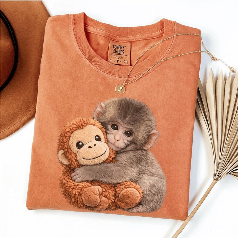 Puede incluir: Camiseta naranja Comfort Colors con un gr&aacute;fico de un beb&eacute; mono abrazando a un mono de peluche. Un collar de oro con un colgante est&aacute; drapeado sobre la camisa. La camiseta tiene la etiqueta Comfort Colors.