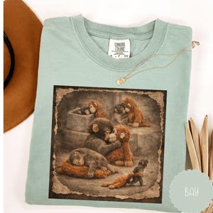 May include: A pale green Comfort Colors t-shirt with a sepia-toned image of monkeys. The image, framed with a distressed border, shows monkeys in different poses. A gold necklace is draped on the shirt.