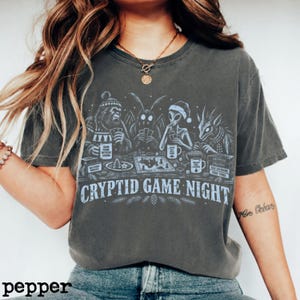 May include: A charcoal gray t-shirt featuring a graphic of Bigfoot, Mothman, an alien, and a creature playing a board game. The text reads "CRYPTID GAME NIGHT" with various small items on the table including mugs labeled "GAME GAME DIFFERENT LEGEND" and "EARTH IS A WILD RIDE".