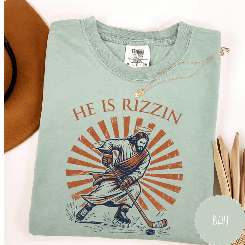 May include: A light green t-shirt with the text "HE IS RIZZIN" above a graphic of Jesus playing hockey. The graphic features Jesus with a halo, wearing a robe and hockey gear, set against a sunburst design. A gold necklace is draped on the shirt.