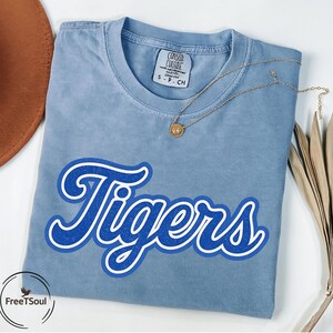 May include: A light blue t-shirt with the word "Tigers" in a blue and white design. A gold necklace with a pendant rests on the shirt. The shirt is folded and has a tag that says "Consed Culari".